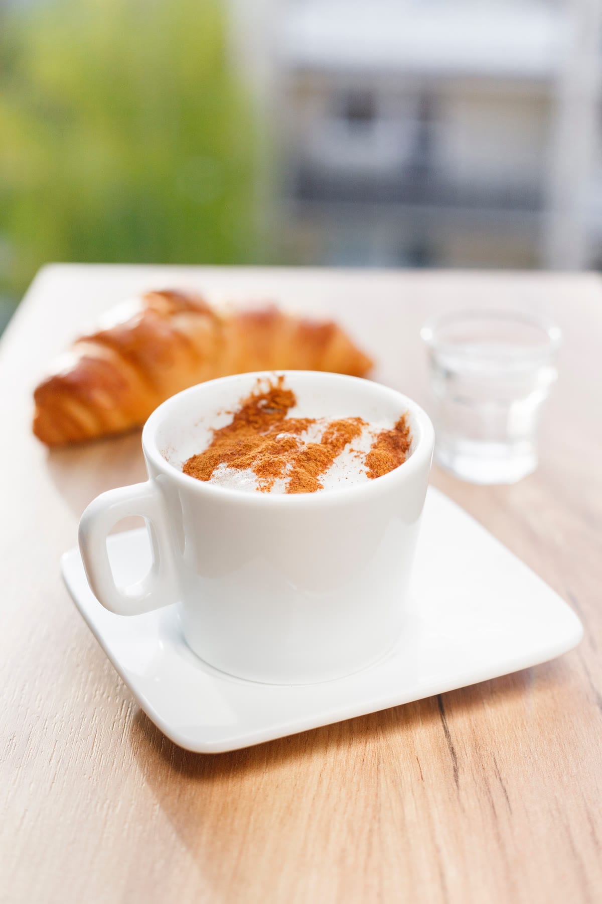 Un cappuccino con il cacao in tazza bianca e un cornetto, su un banco di legno chiaro.