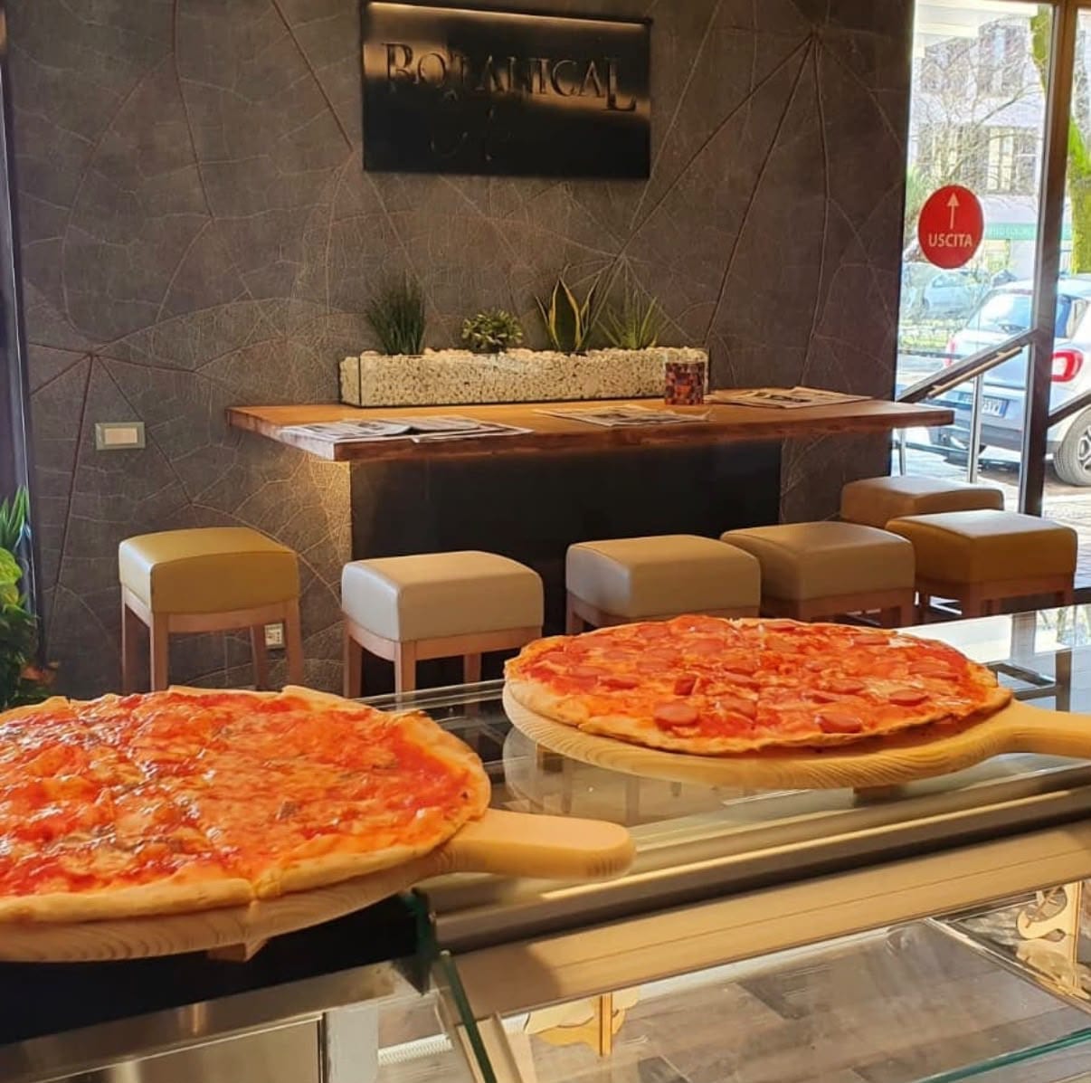 Due pizze sulle pale di legno, sopra la vetrina del banco: una al pomodoro e una al salame piccante. Dietro, la parete con il disegno delle foglie, l'insegna Botanical e gli sgabelli.